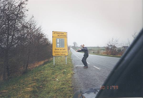 Man poses beside the yellow drive-on-left sign along the road