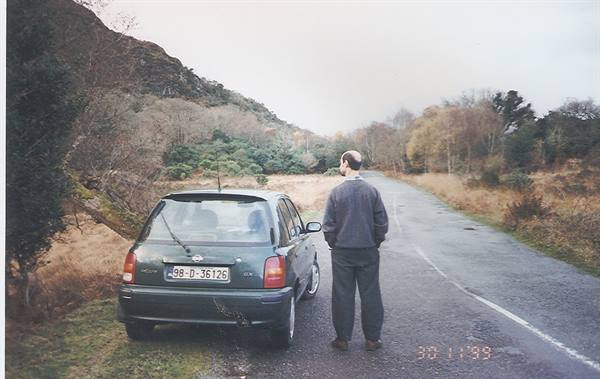 Man standing beside a green car parked on a quiet tree-lined country road