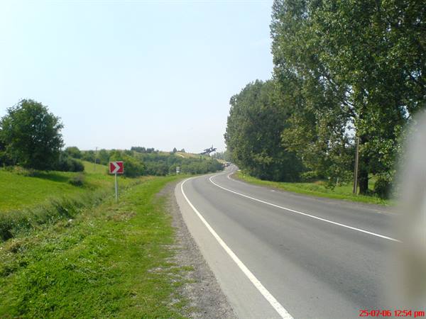 Curving asphalt road with red chevron sign, tall trees and green fields