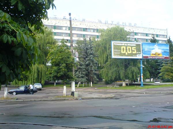 Man next to parked car on urban street with billboards and trees, large Soviet-style building behind