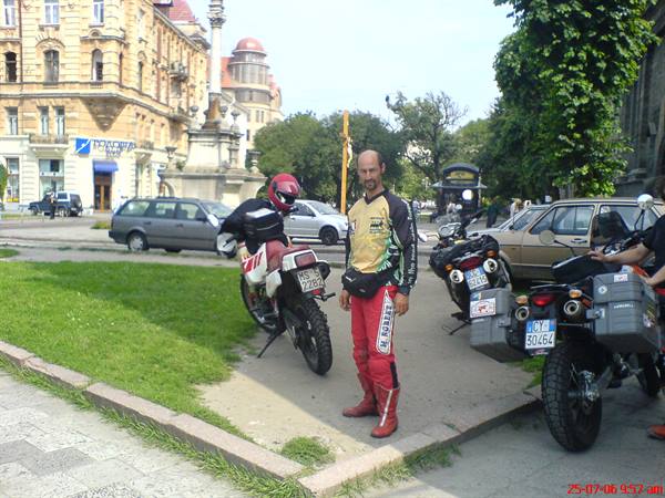 Motorcyclist in red suit beside parked motorcycles in a square with historic buildings