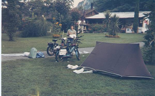 Camper pitches a brown tent beside two loaded motorcycles on a campsite lawn