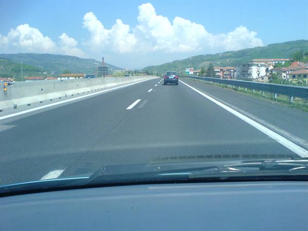 Highway among hills, car ahead, houses and green hills under cloudy sky
