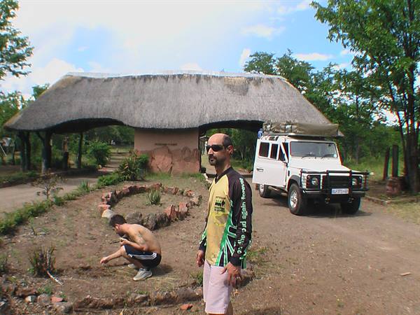 Man in yellow-green shirt in front of thatched-roof lodge and white 4x4