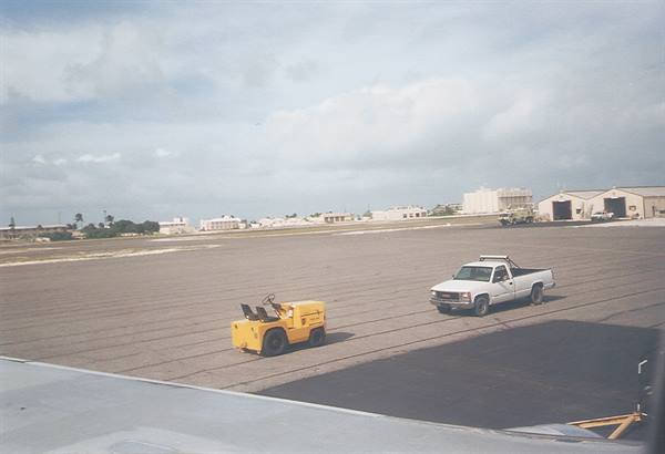 Yellow tug and white pickup on asphalt tarmac, hangars under cloudy sky