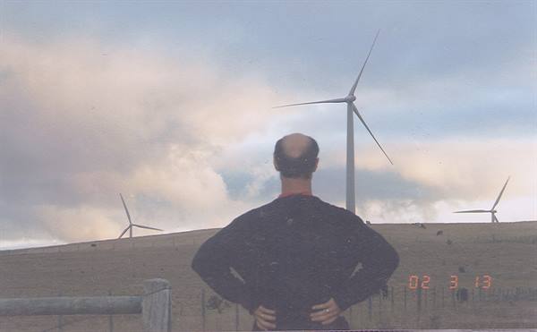 Man in dark sweater watches wind turbines on grassy hill, cloudy sky