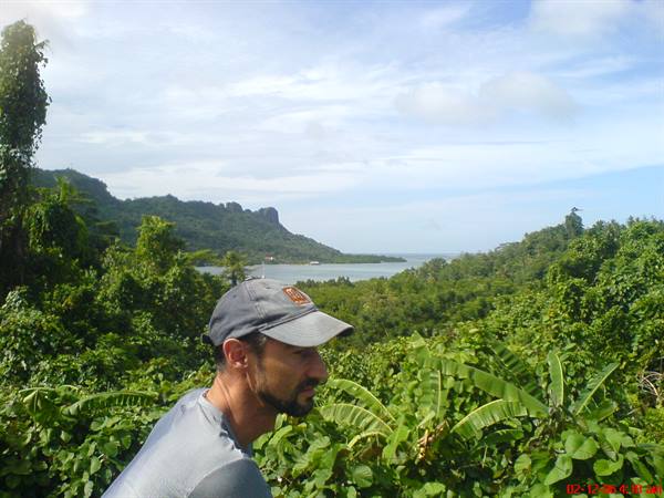 Man in grey cap among banana plants and tropical greenery, green bay between hills