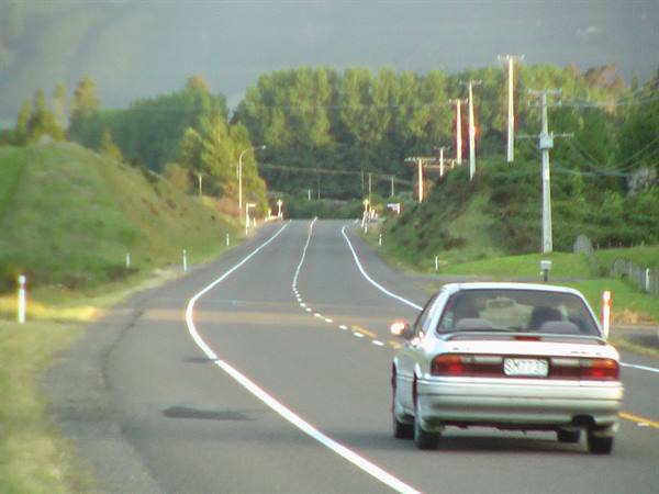 Grey car driving on a rural asphalt road between grassy hills and trees