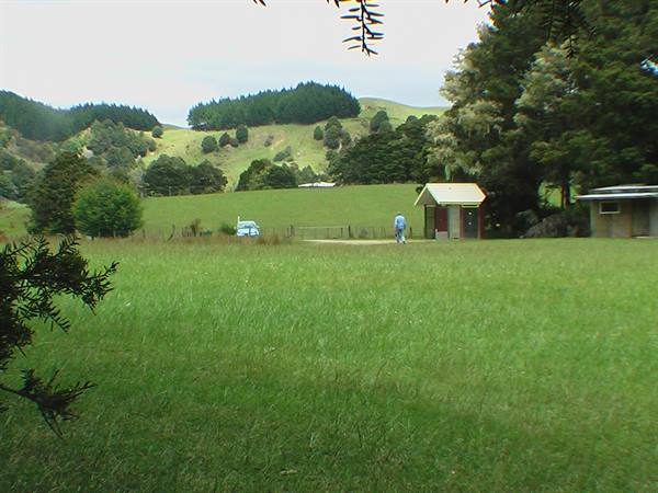 Person walks toward a small building by a green field with wooded hills