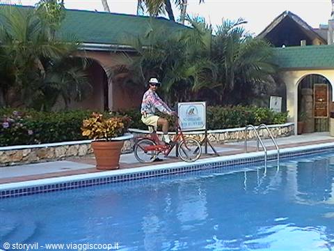 Man riding a bicycle beside a resort pool surrounded by palms