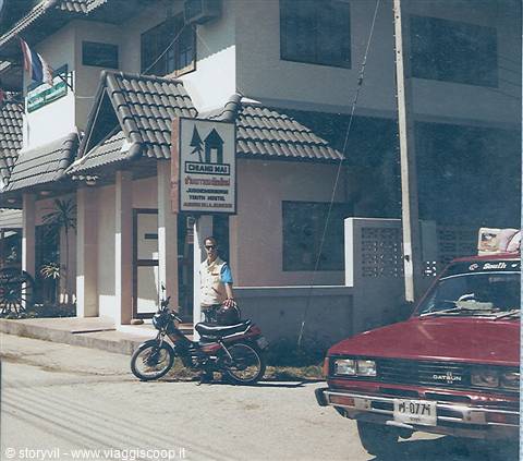 Man beside a motorcycle outside Chiang Mai Guest House, red Datsun car parked nearby