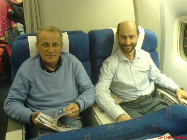 Two men sitting on blue airplane seats, one holding a magazine