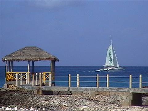 Thatched shelter on stone pier, catamaran with Heineken sail on blue sea