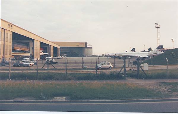 Hangars and parked planes behind the airport fence, a Fiat 500 on the road