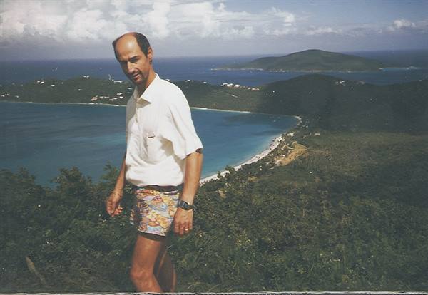 Man in white shirt and floral shorts on green hillside above a turquoise bay