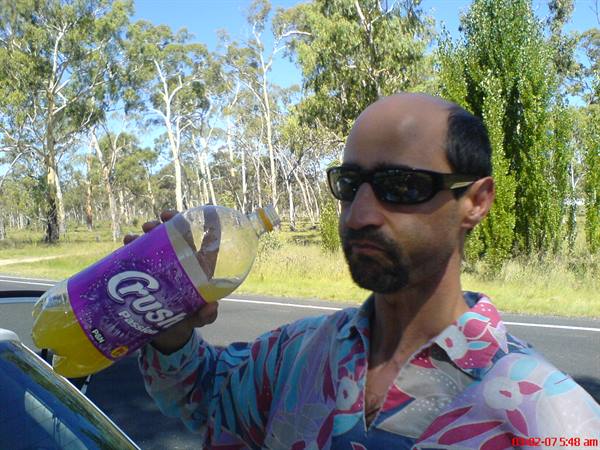 Man in sunglasses raises a Crush bottle beside a road lined with eucalyptus trees