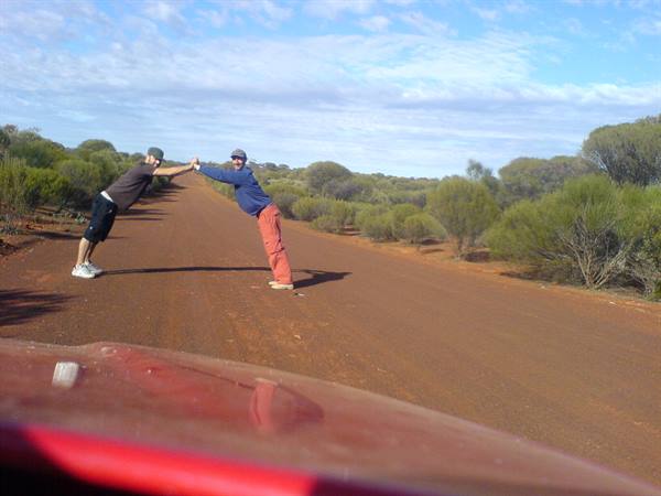 Two guys holding hands on a red dirt road among bushes, red car hood in foreground