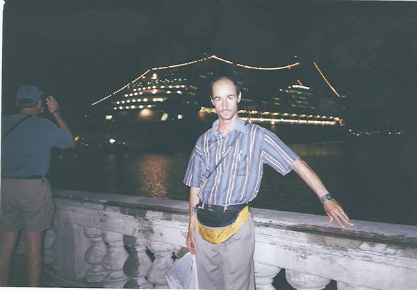 Man with yellow fanny pack by stone balustrade, illuminated cruise ship at night