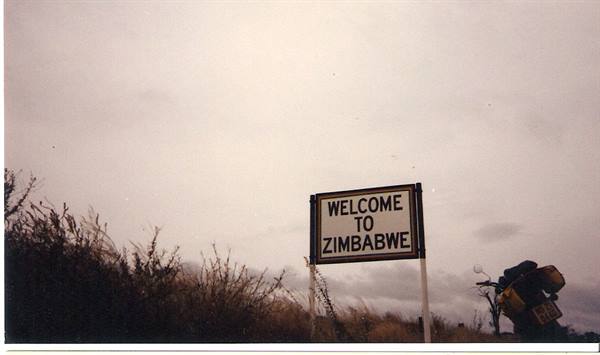 Welcome to Zimbabwe sign among dry bushes, grey sky, motorcycle parked nearby