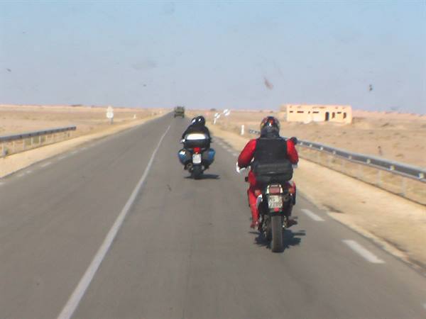 Two motorcyclists with panniers ride a straight desert road, a brick building ahead