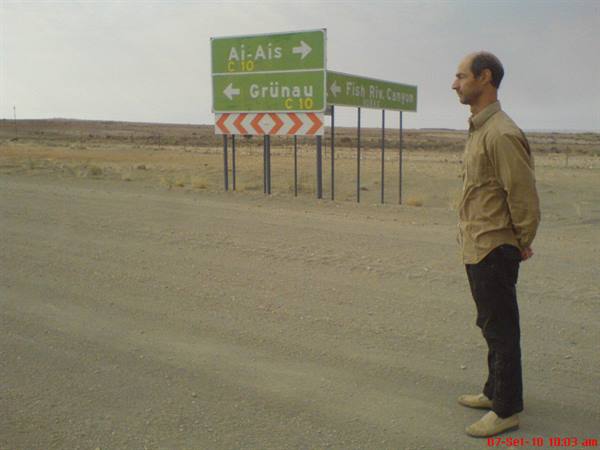 Man in beige shirt beside road signs to Ai-Ais and Grünau in the desert