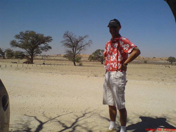 Tourist in red floral shirt and cap standing on sand among dry trees