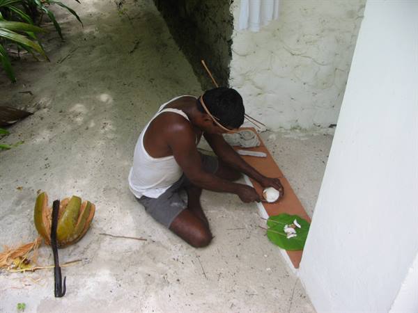 Man kneeling on sand grates a coconut beside a white wall