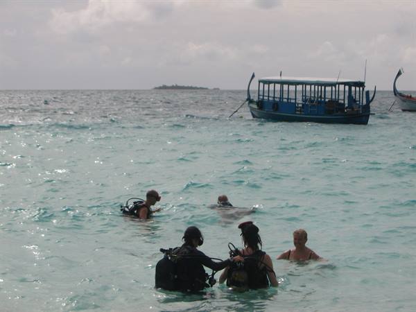 Divers with scuba tanks in turquoise water, blue dhoni boats anchored nearby