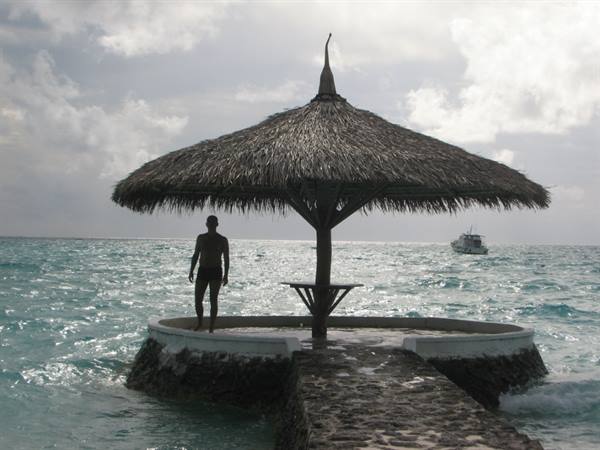 Man silhouetted under a thatched gazebo on a stone platform in turquoise sea