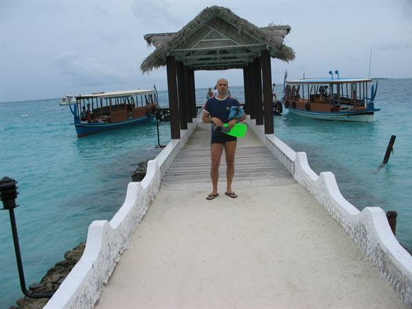 Man in swim trunks holding green fins on thatched jetty, dhoni boats moored beside