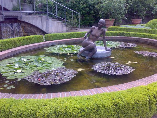 Bronze nude woman statue on stone among water lilies, bordered by box hedges