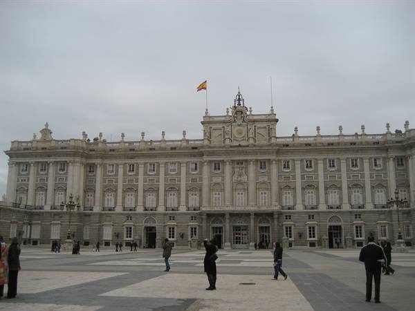 Tourists photograph the Royal Palace of Madrid facade under grey sky
