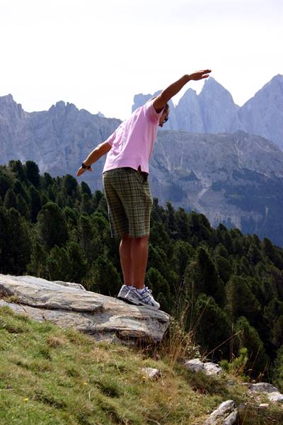 Man in pink shirt balancing on a rock, firs and Dolomites behind