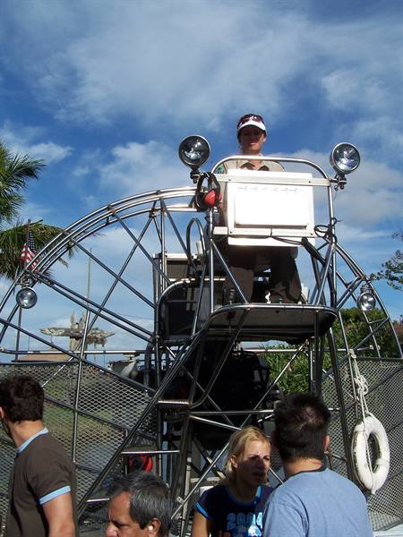 Pilot at the cage of an airboat with fan propeller, tourists nearby under blue sky
