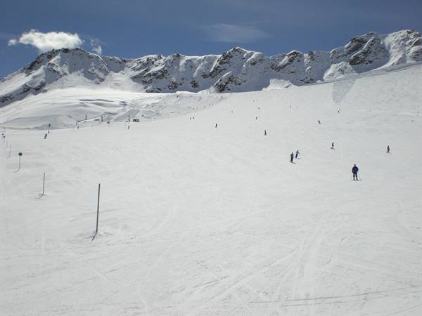 Skiers on wide snowy slope between alpine peaks under blue sky