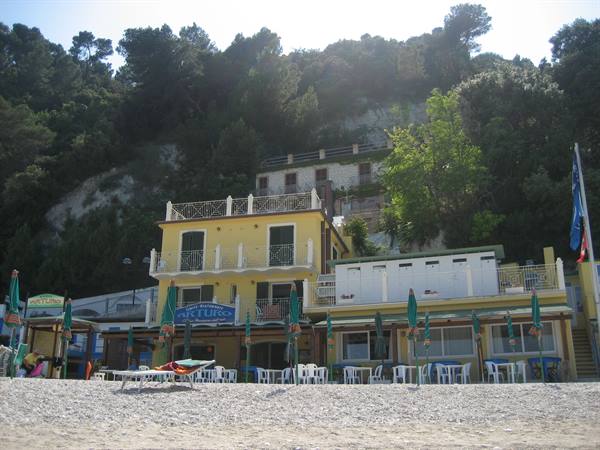 Beach resort Arturo on pebble shore, closed green umbrellas, wooded hillside behind