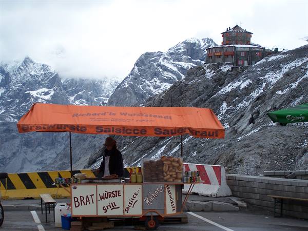 Sausage stand under orange awning among snowy peaks, mountain hut above
