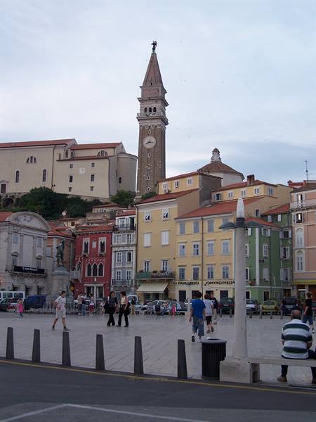 Leaning bell tower of Piran above colorful houses and a square with passersby