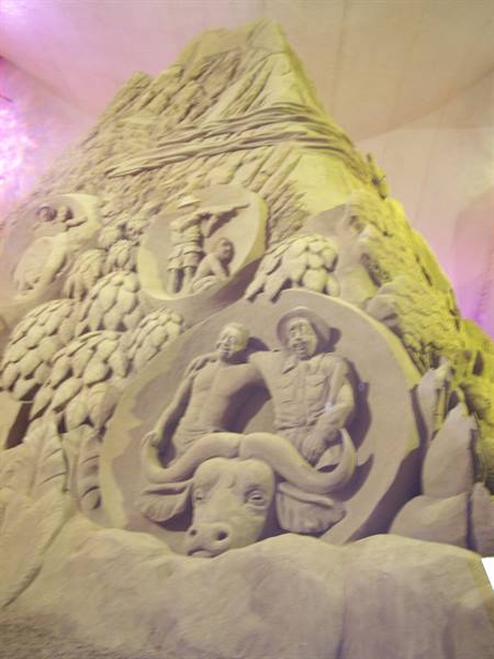 Sand sculpture with human figures and a buffalo, lit by warm lights