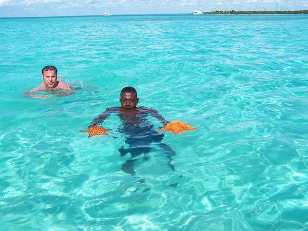 Two swimmers hold orange starfish in clear turquoise water, boats along the shore