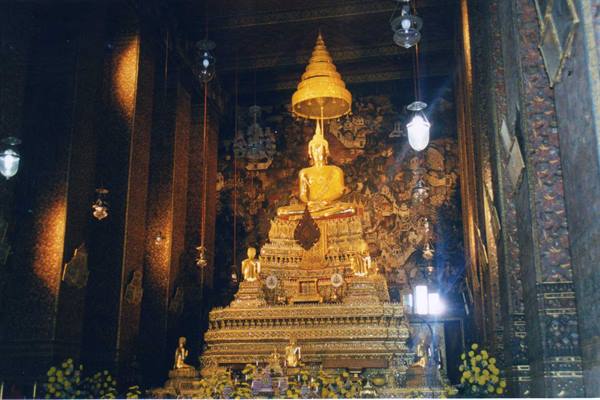 Large seated golden Buddha under ornate umbrella, flower offerings, lit bulbs