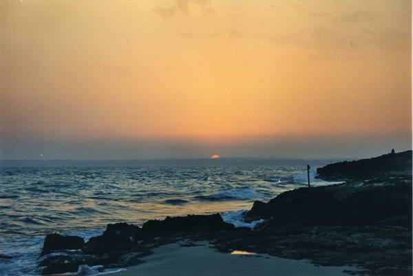 Orange sun sinking over choppy ocean beside black rocks and foam