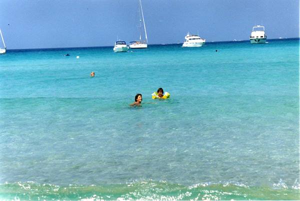 Children swimming in turquoise water, white yachts anchored on the horizon
