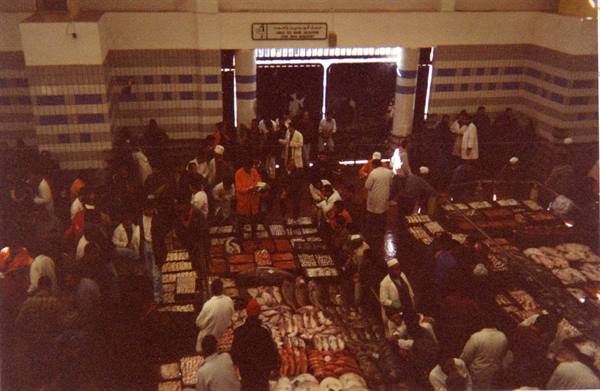 Crowded fish market, stalls with iced fish, people moving, light from entrance