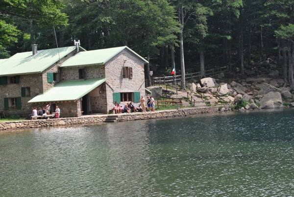 Stone mountain hut with green roof by the lake, people sitting along the shore