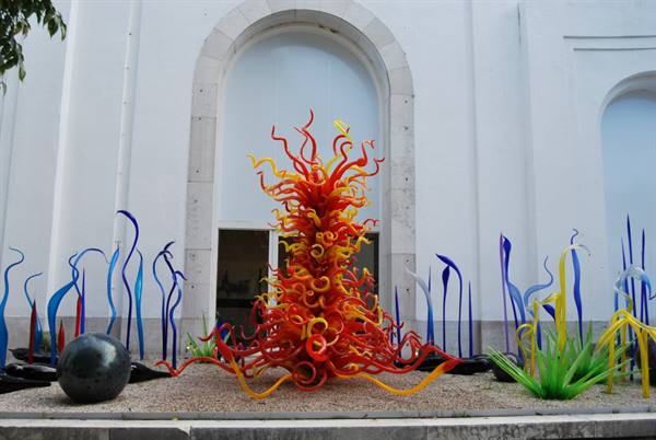 Red and yellow glass sculpture in courtyard before white facade, soft light