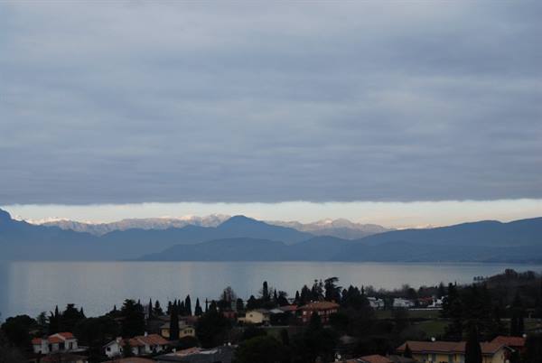 Lake Garda with red rooftops, cypresses and snowy Alps under cloudy sky