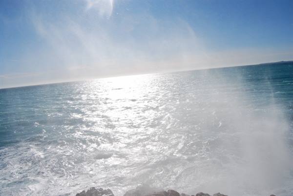 Wind-blown sea spray over rocks, sunlight reflecting on the water