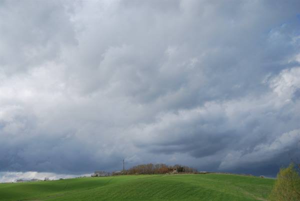 Green fields under grey rain clouds, distant trees and houses on the hill