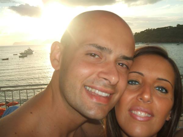 smiling couple close-up, behind them the sea with boats and green hill
